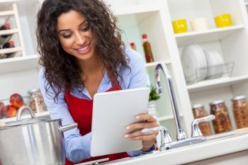 Beautiful girl or young woman looking happy wearing red apron and cooking using tablet computer for social media or recipe in her kitchen at home