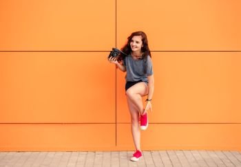 Full Body of trendy girl with vintage camera putting on red sneakers on orange background. Full Body of trendy girl with vintage camera on orange background
