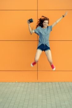 Smiling pretty girl jamping with vintage camera in front of orange wall, blurry image. Pretty girl jamping with vintage camera in front of orange wall
