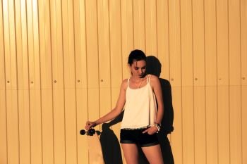 Girl in denim shorts and tank top standing with skateboard in front of yellow mwtal fence and looking down a lot of space for text. Girl in denim shorts and tank top standing with skateboard in front of yellow mwtal fence and looking down