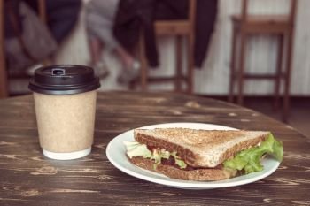 Triangle sandwich and takeaway coffee cup of craft paper on wooden table, with sitting people on background. Triangle sandwich and takeaway coffee cup of craft paper on wooden table