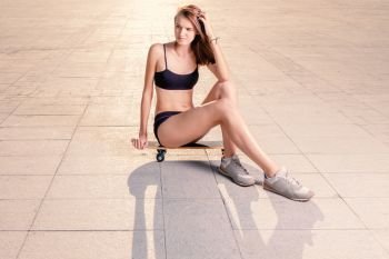 Slim sporty girl is sitting on skateboard in fitness wear and sneakers backlit, a lot of space for text. Slim sporty girl is sitting on skateboard in fitness wear and sneakers backlit, copyspace