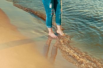 Legs of barefoot girl in ripped jeans on the edge of water on beach in sunset time