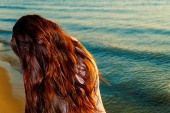 Girl with long red hair on the beach with her face hidden by hair, shot with copyspace on sea water
