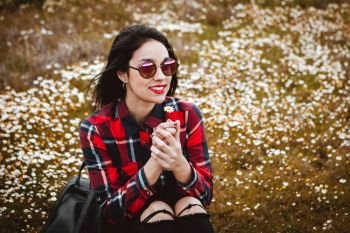 Beautiful woman with sunglasses sitting in the middle of a flowery meadow smelling a daisy
