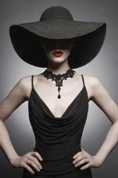 Fashion portrait of young sexy lady with beautiful black hat and evening dress. Stylish elegant woman with modern jewelry. Studio photo of pretty model on grey background.