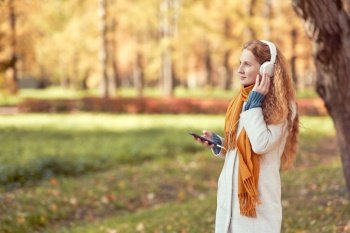 A young red-haired woman in a white coat walks in an autumn park. A girl listens to music in the autumn forest