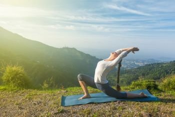 Sporty fit woman practices yoga Anjaneyasana - low crescent lunge pose outdoors in mountains in morning. Woman does yoga asana Anjaneyasana in mountains