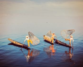 Myanmar travel attraction - Traditional Burmese fishermen balancing with their fishing net on boats at Inle lake in Myanmar famous for their distinctive one legged rowing style. Vintage filtered retro effect hipster style image.  Traditional Burmese fisherman at Inle lake, Myanmar
