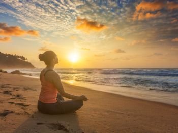 Woman doing yoga - meditate and relax in Padmasana Lotus asana pose with chin mudra outdoors at tropical beach on sunset with dramatic sun. Woman doing yoga oudoors at beach - Padmasana lotus pose