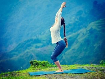 Young sporty fit woman doing yoga asana Utkatasana (chair pose) outdoors in mountains Himalayas in India. Vintage retro effect filtered hipster style image.. Woman doing yoga asana Utkatasana outdoors