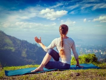 Hatha yoga outdoors - sporty fit woman doing yoga asana Parivrtta Marichyasana (or ardha matsyendrasana) -  seated spinal twist outdoors in mountains in the  morning. Vintage retro effect filtered hipster style image.. Woman practices yoga asana outdoors