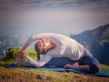 Yoga outdoors - young sporty fit woman doing Hatha Yoga asana parivritta janu sirsasana - Revolved Head-to-Knee Pose - in mountains in the morning. Vintage retro effect filtered hipster style image.. Young sporty fit woman doing Hatha Yoga asana