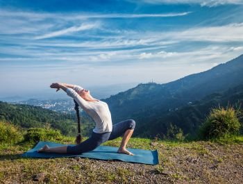 Sporty fit woman practices yoga Anjaneyasana - low crescent lunge pose outdoors in mountains in morning. Woman practices yoga asana Anjaneyasana outdoors