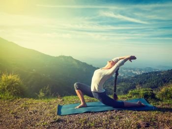 Vintage retro effect hipster style image of sporty fit woman practices yoga Anjaneyasana - low crescent lunge pose outdoors in mountains in morning. Sporty fit woman practices yoga Anjaneyasana in mountains