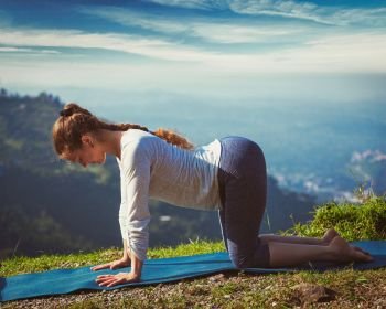 Vintage retro effect hipster style image of sporty fit woman practices yoga asana bitilasana - cow pose outdoors in mountains. Sporty fit woman practices yoga asana bitilasana outdoors