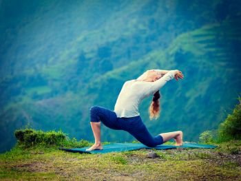 Yoga outdoors - sporty fit woman practices Hatha yoga asana Anjaneyasana - low crescent lunge pose posture outdoors in Himalayas mountains. Vintage retro effect filtered hipster style image.. Sporty fit woman practices yoga asana Anjaneyasana in mountains