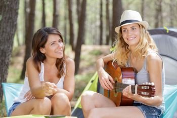young woman playing guitar in the campsite while friend listening