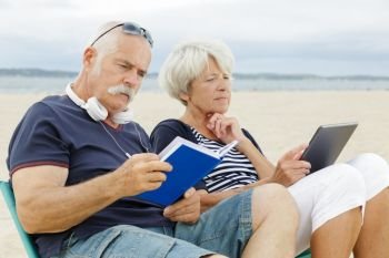 retired couple reading book and tablet on the beach