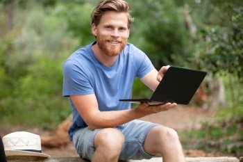 man sits on tree trunk in forest using laptop computer