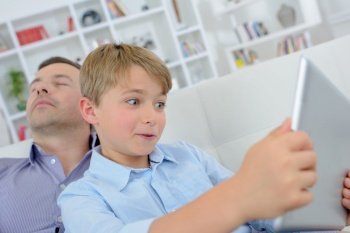 Child wide eyed looking at tablet, father asleep