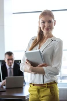 Portrait of confident young businesswoman holding tablet PC with businessman in background at office