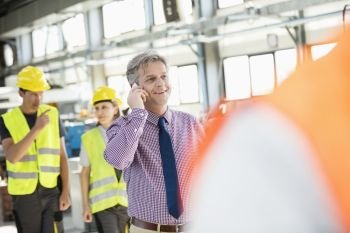 Male supervisor talking on mobile phone with workers in background at industry