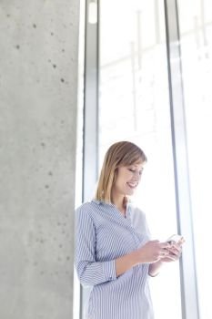 Smiling businesswoman texting on mobile phone by window at office