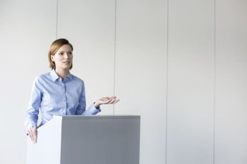 Confident businesswoman giving speech at podium against wall in office
