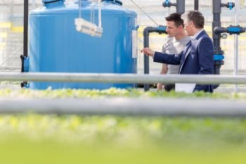 Male botanist and businessman discussing over storage tank in greenhouse