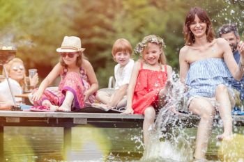 Smiling woman and daughter splashing water in lake while sitting on pier with family during summer picnic