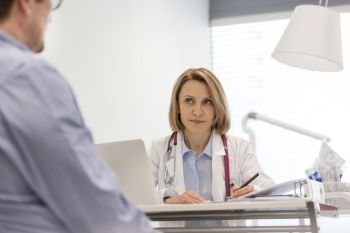 Doctor discussing with mature patient while writing prescription at desk in hospital