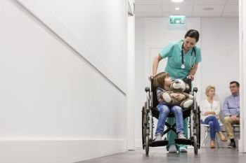 Nurse pushing boy with teddybear on wheelchair while patients waiting in hospital corridor