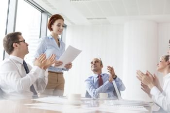 Colleagues applauding businesswoman standing with document in boardroom at office during meeting