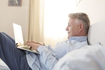 Side view of smiling mature man using laptop while lying on bed at home