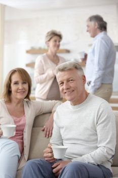Portrait of smiling mature couple holding coffee while sitting against friends at home