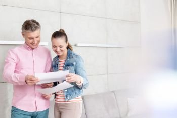 Smiling couple reading agreement paper while standing against wall in apartment
