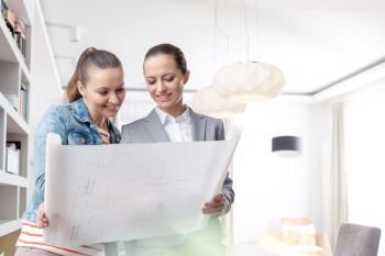 Smiling young saleswoman and female client looking at blueprint in apartment
