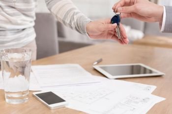 Hand of saleswoman giving house keys to female client over documents on table in apartment