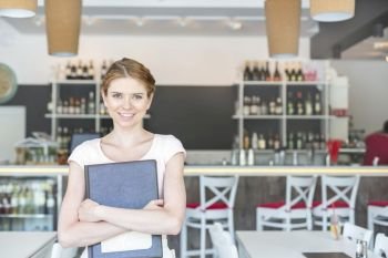 Portrait of smiling young waitress holding menu while standing at restaurant