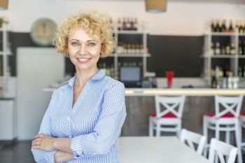 Portrait of confident blond owner standing with arms crossed at restaurant