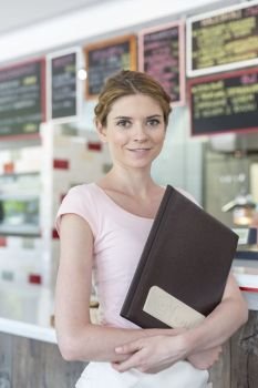 Portrait of smiling young waitress holding menu while standing against counter at restaurant