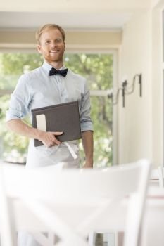 Portrait of confident young waiter holding menu while standing at restaurant