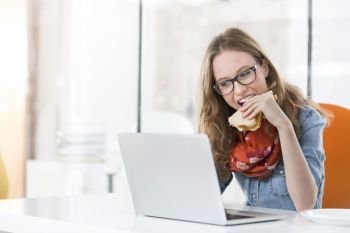 Beautiful businesswoman eating sandwich while using laptop in office