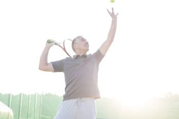 Back lit of confident mature man serving on tennis court against clear sky