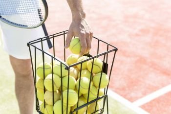 Midsection of man picking tennis ball from metallic basket on sunny day