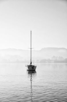 Beautiful unplugged landscape image of sailing yacht sitting still in calm lake water in Lake District during peaceful misty Autumn Fall sunrise