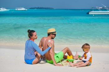Family on beach, young couple with three year old boy. Woman applying sun screen protection lotion on man’s back. Summer vacation at Maldives.