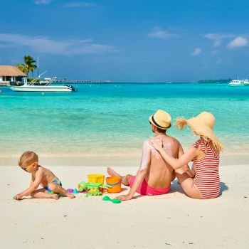 Family on beach, young couple with three year old boy. Woman applying sun screen protection lotion on man’s back. Summer vacation at Maldives.