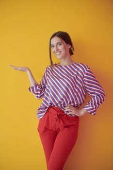 Portrait of happy smiling young beautiful woman in a presenting gesture with open palm isolated on yellow background. Female model in modern fashionable clothes posing in the studio